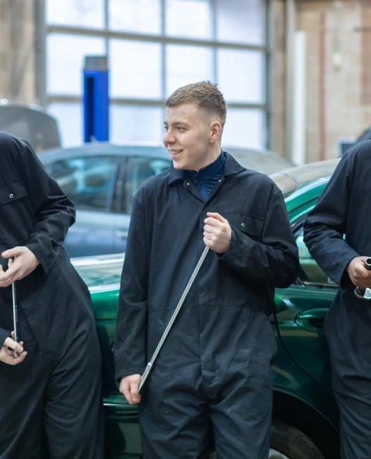 Three students in black overalls laughing and holding tools while standing in front of a green car in the workshop. Three students in black overalls laughing and holding tools while standing in front of a green car in the workshop.